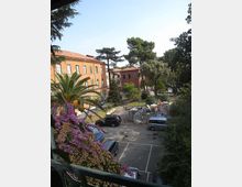 Veduta dall’alto di un cortile con parcheggio e aiuole, circondato da edifici in mattoni dai tetti in tegole. Sono presenti alberi alti, una palma e fioriture colorate, oltre ad alcune aree con materiali e strutture da cantiere.
