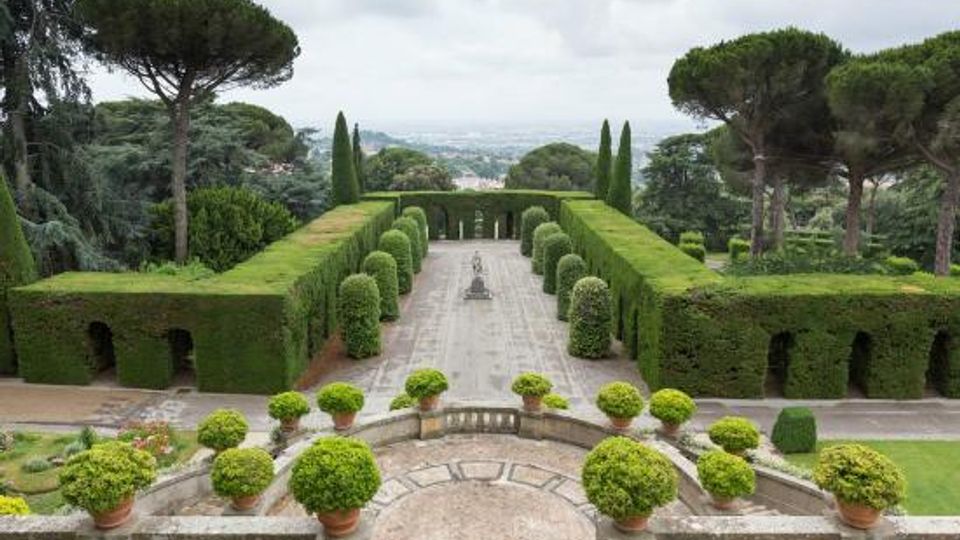 Giardino formale con viali simmetrici delimitati da alte siepi potate e filari di cipressi, con una fontana centrale su pavimentazione in pietra. In primo piano una terrazza con balaustra e vasi di arbusti sferici, circondata da grandi pini e panorama collinare sullo sfondo.