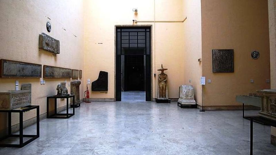 Sala museale con pavimento in pietra chiara e alte pareti color crema, con lapidi e reperti archeologici esposti su supporti e lungo le pareti. Al centro si apre un grande portale scuro che conduce a un ambiente interno più buio.
