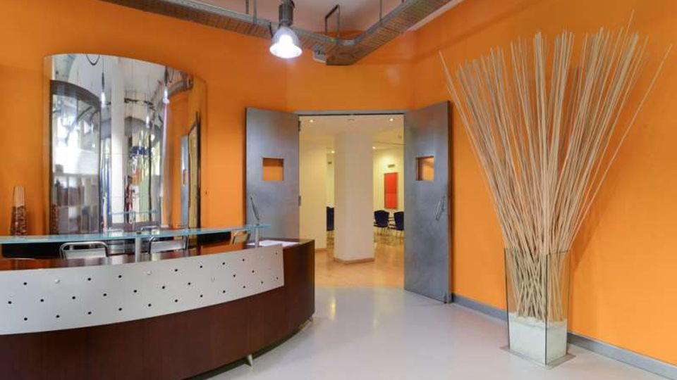 Atrio interno moderno con pareti arancioni, pavimento chiaro e soffitto con canaline e lampade a vista. In primo piano c’è un banco reception curvo con pannello traforato e specchio, mentre sul lato destro si vede un grande vaso con canne decorative e una porta doppia che conduce a un corridoio.