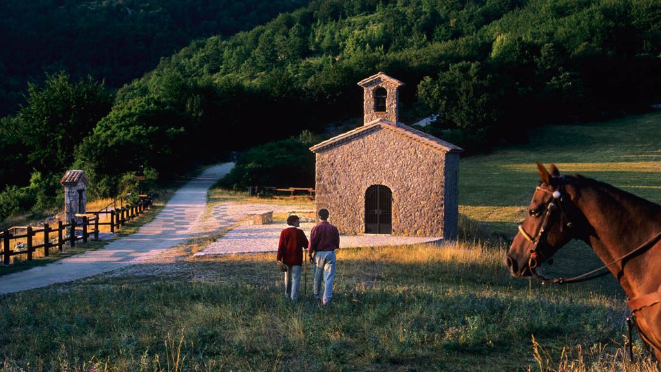 Piccola chiesa rurale in pietra con campanile a vela e portale ad arco, situata in un prato lungo una strada sterrata con staccionate. Sullo sfondo, colline ricoperte di bosco e campi aperti.