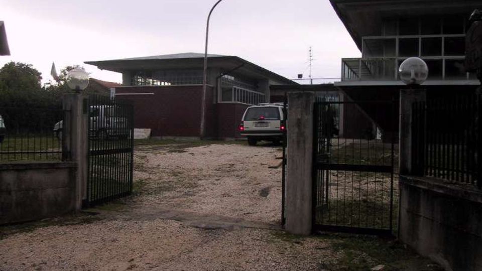Ingresso con cancello metallico aperto su un vialetto sterrato che conduce a un complesso di edifici bassi in stile industriale, con ampie finestre e tetti aggettanti. Nell’area interna si vedono recinzioni, pilastri in cemento con lampade sferiche e un lampione stradale al centro.