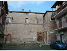 Piazzetta urbana con un grande edificio in muratura intonacata e invecchiata, con finestre strette e portali in pietra, affiancato da palazzi con balconi e ringhiere in metallo. In primo piano si vedono la pavimentazione, un’auto parcheggiata e lampioni a parete sotto un cielo sereno.