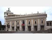 Facciata di un grande palazzo neoclassico con due livelli di arcate e finestre, decorazioni in pietra e statue sul cornicione, affacciato su un’ampia piazza lastricata con lampioni. Sul tetto spiccano sculture e una quadriga, mentre a lato si vede una torre angolare.