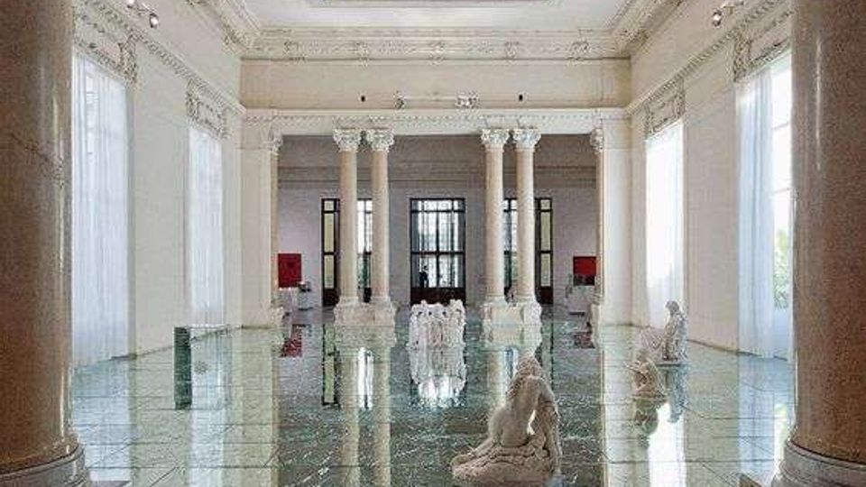Ampia sala interna in stile neoclassico con alte colonne, grandi finestre con tende chiare e soffitto a cassettoni con lucernario centrale. Pavimento lucido in marmo venato con piccole sculture bianche disposte nello spazio e riflessi sull’acqua o su una superficie molto brillante.