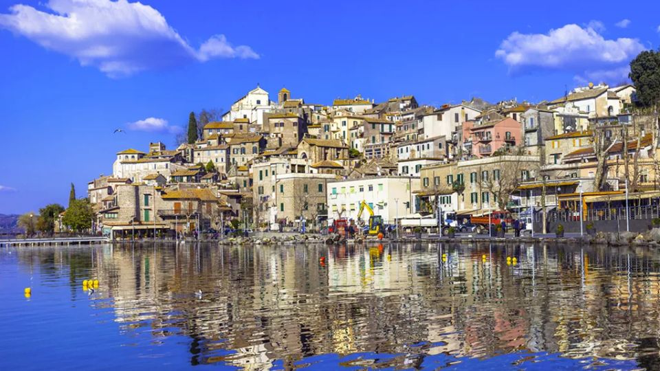 Borgo collinare con case in pietra e facciate chiare addossate al pendio, affacciato su un lungolago con piccole strutture portuali. In primo piano l’acqua calma riflette gli edifici e il cielo azzurro con alcune nuvole.