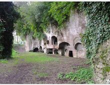 Parete rocciosa lunga con numerose nicchie e aperture ad arco scavate nella pietra, alcune con ingressi rettangolari, lungo un sentiero di terra. La scarpata è ricoperta di edera e vegetazione, con alberi e prato ai lati.