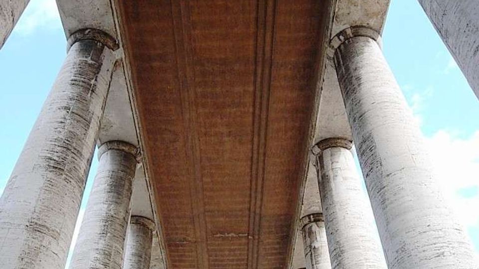 Vista dal basso di un viadotto in cemento armato, con l’intradosso marrone della carreggiata che corre in prospettiva tra alte colonne cilindriche. Il cielo azzurro è visibile ai lati, evidenziando la struttura ripetuta dei piloni.