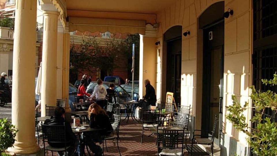 Portico di un edificio con grandi colonne chiare e soffitto alto, affacciato su una strada urbana. Sotto il portico c’è un’area esterna di caffè con tavolini e sedie in metallo, porte ad arco e alcune piante in vaso lungo il lato.