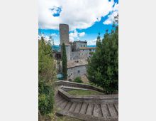 Scalinata in pietra con muretto curvo che scende verso un borgo medievale in muratura, con edifici dai tetti in coppi e una torre rettangolare alta. Vegetazione ai lati della scala e panorama di colline sullo sfondo sotto un cielo con nuvole.