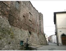 Muro esterno di una fortificazione in pietra e mattoni con piccole feritoie e finestre grate, affiancato da una strada urbana. In primo piano si vede una piccola fontana in pietra ai piedi della muratura, con lampioni e edifici moderni sul lato opposto.