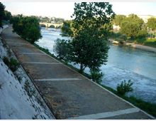 Passeggiata lungo un fiume con ampio argine in cemento e sentiero in ghiaia sul lato sinistro, affiancato da alberi e vegetazione. In lontananza si vede un ponte ad archi che attraversa il fiume, con la città sullo sfondo.