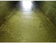 Interno di un sottopassaggio pedonale in cemento con pareti laterali ondulate e pavimento in mattonelle, illuminato da luce intensa all’uscita in fondo al tunnel. L’ambiente appare lungo e stretto, con segni di sporco e umidità sul pavimento.