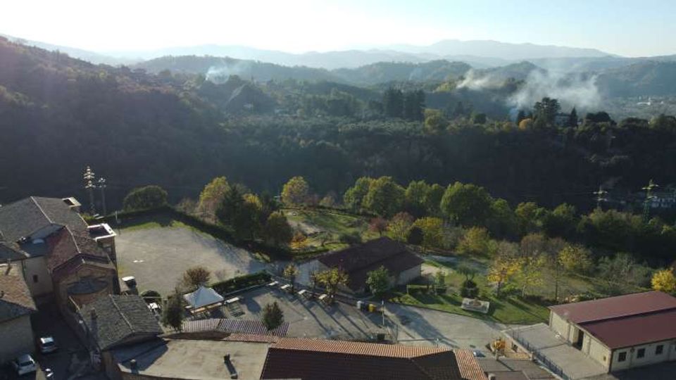 Veduta panoramica dall’alto di un piccolo centro con edifici dai tetti in laterizio, un piazzale e aree verdi alberate. Sullo sfondo si estendono colline e montagne boscose con leggere velature di nebbia o fumo tra le valli.