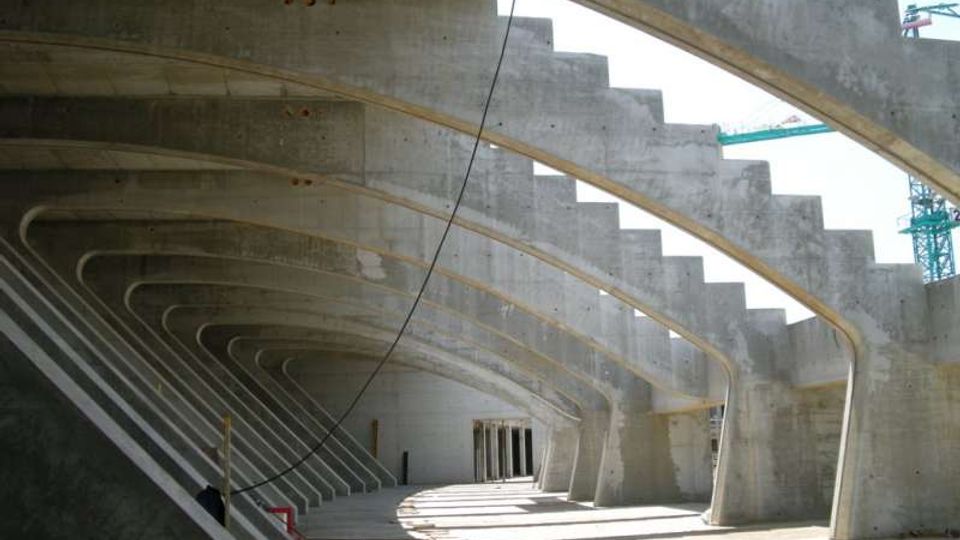 Interno di una grande struttura in cemento armato con una serie di archi e travi curve ripetute che formano un corridoio coperto. Sulla destra si vede una scalinata integrata nella parete e, sullo sfondo, aperture verso l’esterno con luce naturale.