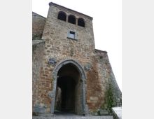 Portale d’ingresso in pietra di una struttura storica fortificata, con grande arco a tutto sesto che introduce a un passaggio coperto. Sopra l’arco si vede una torretta in muratura con tre arcate aperte e un piccolo finestrino, circondata da muri in pietra e pavimentazione in ciottoli.