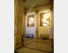 Interno di una galleria o museo in pietra, con colonne e cornici classiche e pavimento a riquadri. Sulla parete sono esposti due grandi bassorilievi illuminati, con panche in pietra e un busto su piedistallo tra le colonne.