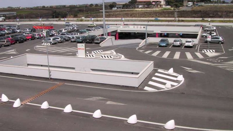 Ampio parcheggio asfaltato con numerose auto, corsie di ingresso e uscita segnalate da strisce pedonali e frecce direzionali. Al centro si vede una rampa di accesso a un parcheggio coperto con barriere e segnaletica, sullo sfondo campagna e colline.