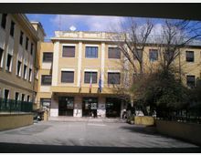 Edificio pubblico a tre piani con facciata gialla e colonne bianche, ingresso centrale con gradini e tre bandiere sopra la pensilina. Davanti si trova un cortile asfaltato, con recinzioni ai lati e alberi spogli sulla destra.