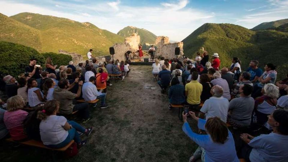 Anfiteatro all’aperto con panche disposte lungo un corridoio centrale, davanti a un piccolo palco tra muri in pietra di un rudere. Sullo sfondo si vedono colline e montagne verdi sotto un cielo sereno.