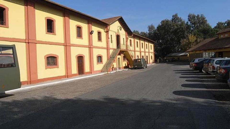 Edificio lungo a due piani in stile industriale, con facciata giallo chiaro e cornici rosse, finestre ad arco e scala esterna centrale che conduce a un ingresso sopraelevato. Davanti si estende un ampio piazzale asfaltato con auto parcheggiate sul lato destro e alberi sullo sfondo sotto un cielo sereno.