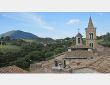 Veduta panoramica di un borgo collinare con tetti in coppi e una chiesa in pietra con campanile e piccola torretta. Sullo sfondo si estendono colline e un monte verde sotto un cielo sereno.