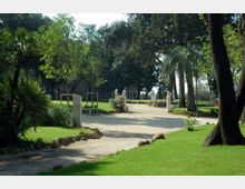 Viale curvilineo in un parco con prato curato e alberi alti, fiancheggiato da palme e statue in pietra su piedistalli. Sentiero chiaro con bordi in pietra e vegetazione ornamentale lungo i lati.