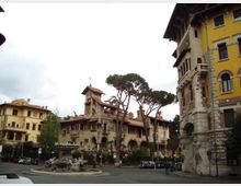 Piazza urbana con rotonda e fontana scultorea al centro, circondata da edifici storici in stile liberty con torrette e logge. Sullo sfondo si vedono alti pini marittimi e facciate in pietra e intonaco, con un palazzo giallo a destra.