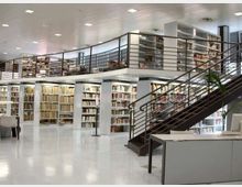 Interno di una biblioteca moderna a due livelli, con ampie scaffalature di libri e un soppalco con ringhiere metalliche. Una scala in metallo collega i piani in un ambiente luminoso con pavimento chiaro e arredi essenziali.