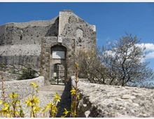 Passerella in pietra con parapetti che conduce a un ingresso ad arco con cancello metallico, inserito in mura di una fortificazione in pietra parzialmente in rovina. Intorno si vedono rocce, cespugli e piccoli alberi, con cielo azzurro sullo sfondo.