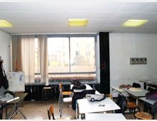 Aula scolastica con banchi e sedie disposti in modo irregolare, soffitto a pannelli con luci fluorescenti e pareti bianche. Sul fondo una grande finestra con tenda parziale affaccia su edifici esterni; presenti uno sgabello e alcuni pannelli/quadri appoggiati ai lati.