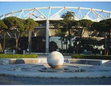 Veduta esterna di un grande stadio moderno con struttura bianca a travi e copertura a membrana, parzialmente nascosto da alberi. In primo piano si vede una piazza con una fontana circolare e una grande sfera in pietra al centro.