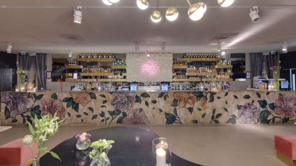 Interno di un bar con lungo bancone frontale decorato da un grande motivo floreale, dietro al quale si trovano scaffali illuminati con bottiglie e bicchieri. Il soffitto è basso con faretti e lampade a sospensione, e in primo piano c’è un tavolo con piccoli vasi di fiori e una candela.