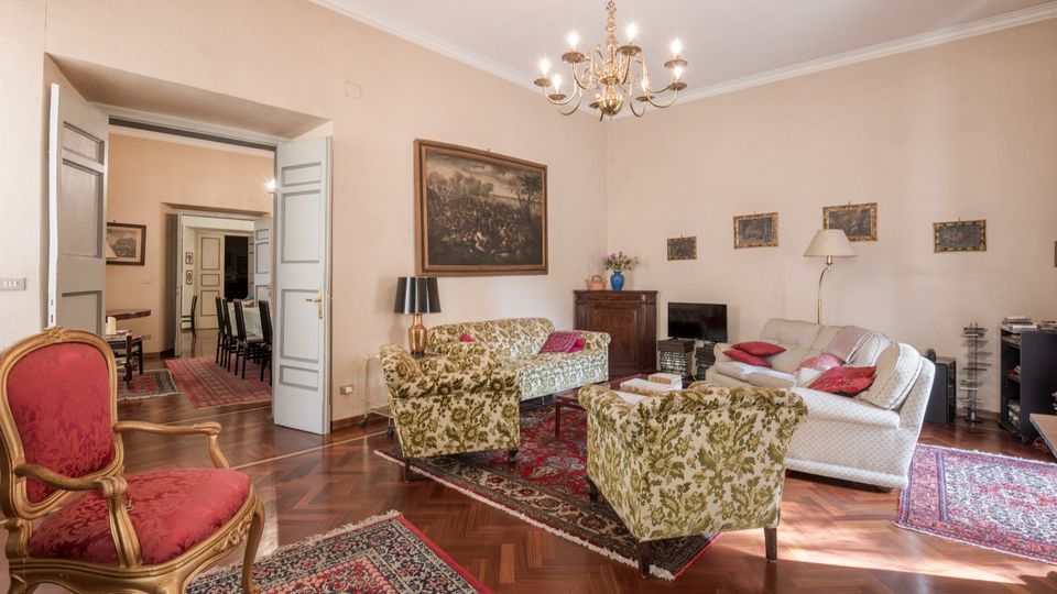 Ampio soggiorno arredato in stile classico con pavimento in parquet, tappeti persiani e salotto composto da divani e poltrone a motivi floreali. Al soffitto è appeso un lampadario in ottone e sulle pareti chiare sono presenti quadri, con una porta doppia che conduce a una sala da pranzo.
