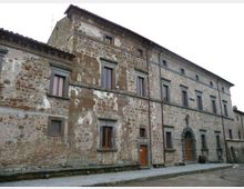 Edificio storico in pietra con facciata irregolare e intonaco consumato, disposto su più piani con finestre rettangolari incorniciate e tetto a falde. Sul lato destro si vede un grande portale ad arco in pietra che si affaccia su una strada lastricata.