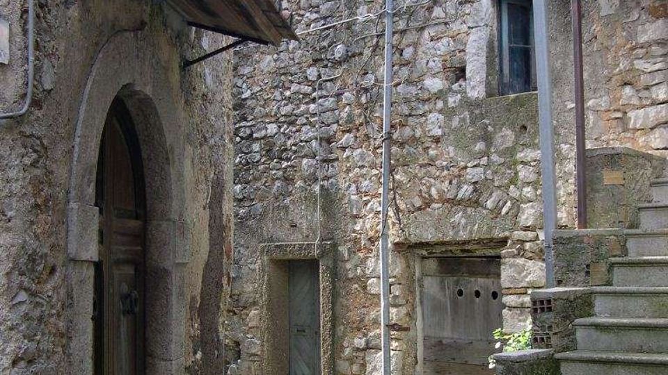 Vicolo stretto in un borgo con edifici in pietra grezza, porte ad arco e piccole finestre, con cavi e tubazioni a vista sulle facciate. A destra una scalinata in pietra sale verso un pianerottolo coperto da una tettoia metallica.