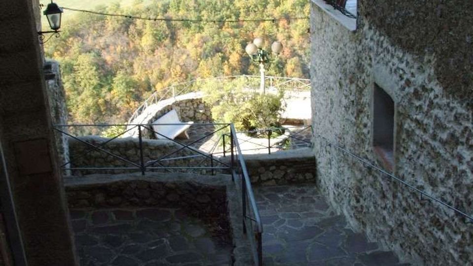 Scalinata e terrazzamento in pietra tra edifici in muratura, con ringhiere metalliche e lampione, affacciati su una valle verde con boschi e campi ondulati. Sullo sfondo si vedono colline boscose che chiudono il panorama.