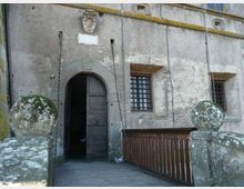 Facciata in pietra di un edificio storico con portale ad arco e grande porta in legno, affiancata da finestre con inferriate. Un breve ponte o passerella in legno con balaustra e pilastri in muratura sormontati da sfere conduce all’ingresso.