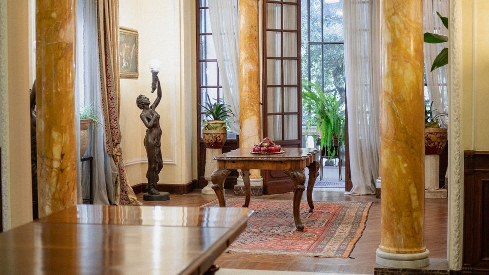 Interno di una sala in stile classico con grandi colonne marmorizzate e arco decorato, pavimento in parquet e tappeti orientali. Al centro un tavolino e, sul fondo, ampie porte a vetri con tende leggere che si aprono su un terrazzo con piante.