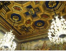 Soffitto interno di una sala storica con cassettonato dorato a riquadri geometrici blu e decorazioni in rilievo, con grandi lampadari in vetro sospesi. Sulle pareti corre un fregio dipinto con scene paesaggistiche e figure, sotto una cornice ornamentale.