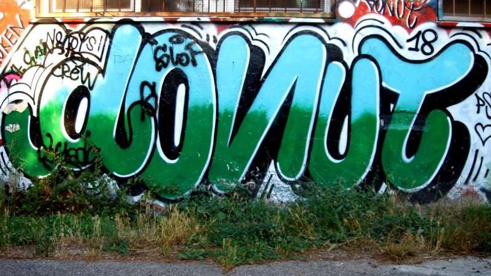 Muro esterno di un edificio con finestre protette da grate, ricoperto da un grande graffito con lettere blu e verdi su sfondo bianco. In primo piano si vede una striscia di asfalto con erba e vegetazione bassa lungo la base del muro.