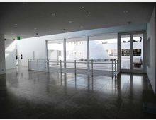 Atrio interno moderno con pavimento in grandi lastre lucide grigie, soffitto con faretti incassati e parapetto in metallo. Ampie vetrate e una porta a vetri si affacciano su una terrazza esterna con elementi architettonici bianchi.