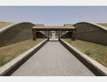 Ingresso a un complesso architettonico con un lungo viale pavimentato in mattoni tra basse aiuole erbose, affiancato da due grandi muri curvi in mattoni. In fondo si vede un portico con tetto in tegole sostenuto da pilastri e l’accesso a un edificio interno.