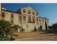 Facciata di un grande edificio storico in pietra e mattoni, con finestre incorniciate, archi e un piccolo balcone centrale. Davanti si apre un cortile con pavimentazione in mattoni, aiuole e alberi in un’area verde.