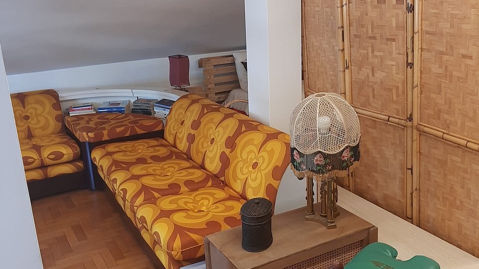 Salottino in mansarda con soffitto inclinato e pavimento in parquet, arredato con divani a fiori arancioni, tavolino basso e libreria. Lungo la parete c’è un paravento in bamboo e un mobiletto con lampada decorativa accanto a uno sgabello verde.