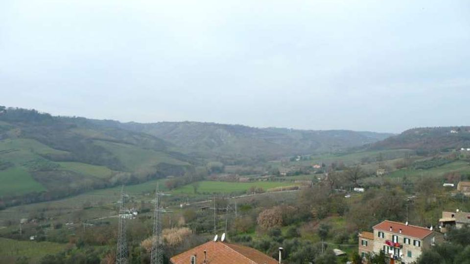 Vista panoramica di una valle collinare con campi verdi e boschetti, sotto un cielo nuvoloso. In primo piano si vedono case con tetti in tegole rosse, una strada e tralicci dell’alta tensione che attraversano il paesaggio.