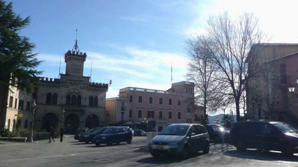 Piazza cittadina con ampio parcheggio e auto, affacciata su un edificio storico in stile municipale con portici ad arcate, merlature e torre centrale. Sul lato destro si vedono alberi spogli e altri edifici a più piani sotto un cielo sereno.