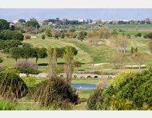Ampio campo da golf con fairway ondulati, alberi sparsi e alcune aree di sabbia, con un piccolo laghetto in primo piano. Sullo sfondo si vedono campi agricoli e un’area urbana lontana all’orizzonte.