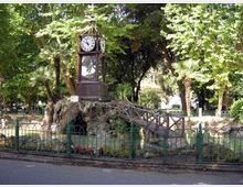 Orologio monumentale su una struttura in pietra simile a una grotta, con una piccola passerella in legno, all’interno di un parco alberato. In primo piano si vede una recinzione in ferro e aiuole fiorite lungo il bordo della strada.