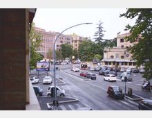 Ampio incrocio urbano con più corsie e parcheggi, con auto e strisce pedonali visibili. Sullo sfondo si vedono edifici multipiano, un edificio basso con finestre ad arco e murales, e alberi tra cui una palma e una conifera.